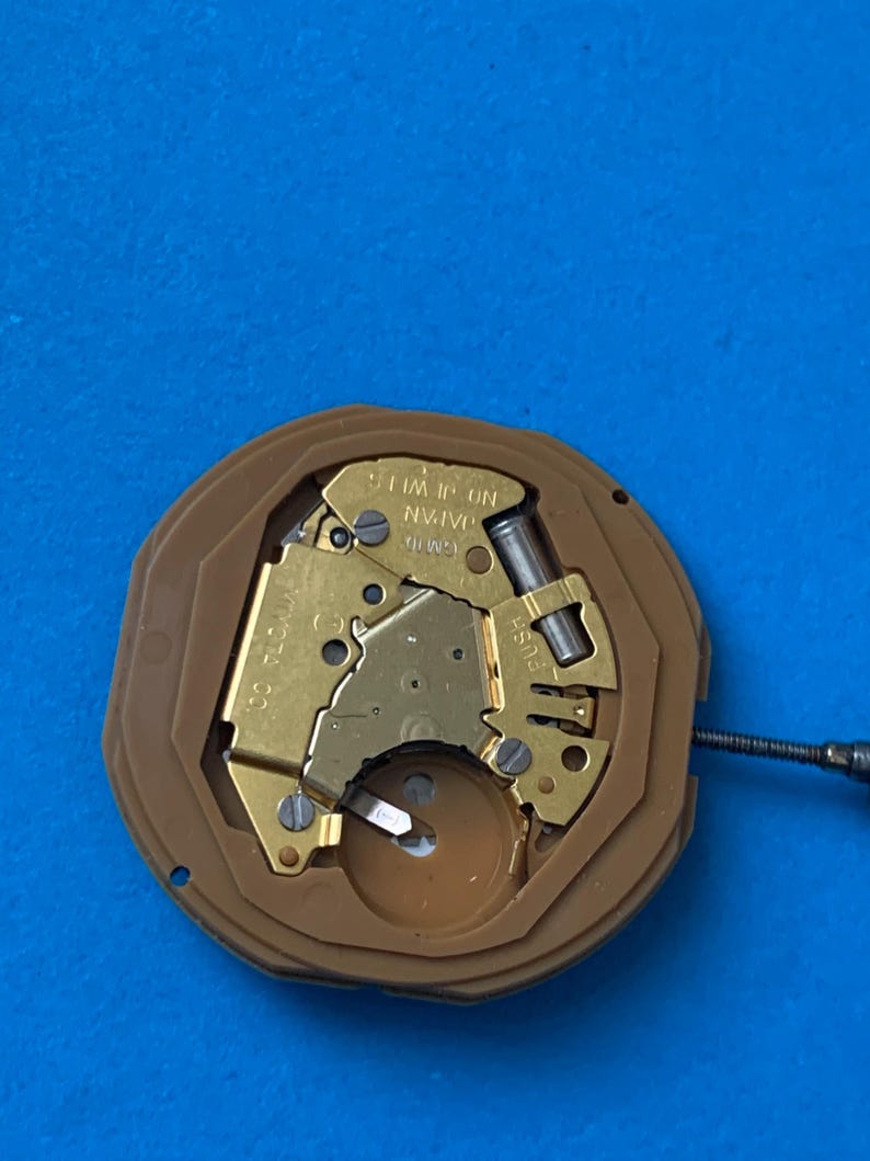 May include: Close-up of a watch movement with gold-colored metal components and a brown plastic casing. The text "JAPAN" and "1 JEWEL" are visible. A small screw is positioned to the right of the movement.