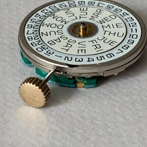 Puede incluir: Primer plano de un movimiento de reloj con una esfera blanca que muestra los d&iacute;as de la semana y n&uacute;meros. La esfera est&aacute; rodeada por una caja de metal plateado. Una corona dorada est&aacute; unida al lateral. El movimiento del reloj est&aacute; sobre una superficie clara.