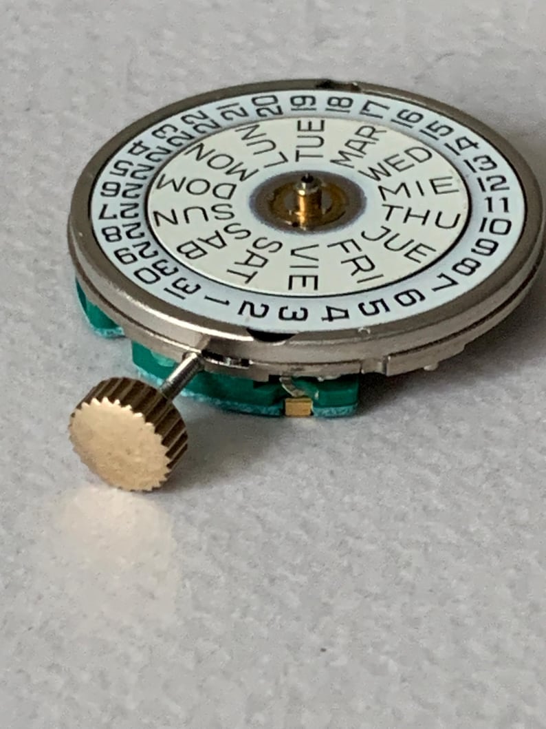 Puede incluir: Primer plano de un movimiento de reloj con una esfera blanca que muestra los d&iacute;as de la semana y n&uacute;meros. La esfera est&aacute; rodeada por una caja de metal plateado. Una corona dorada est&aacute; unida al lateral. El movimiento del reloj est&aacute; sobre una superficie clara.