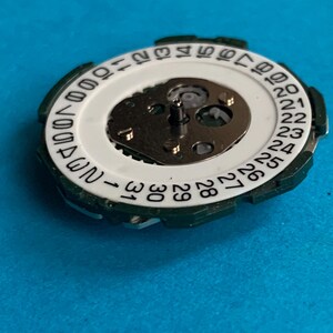 May include: A close-up of a watch movement, showing a white dial with black numbers for the days of the month. It has a green outer ring and a central mechanism with metallic parts. The background is a solid blue.