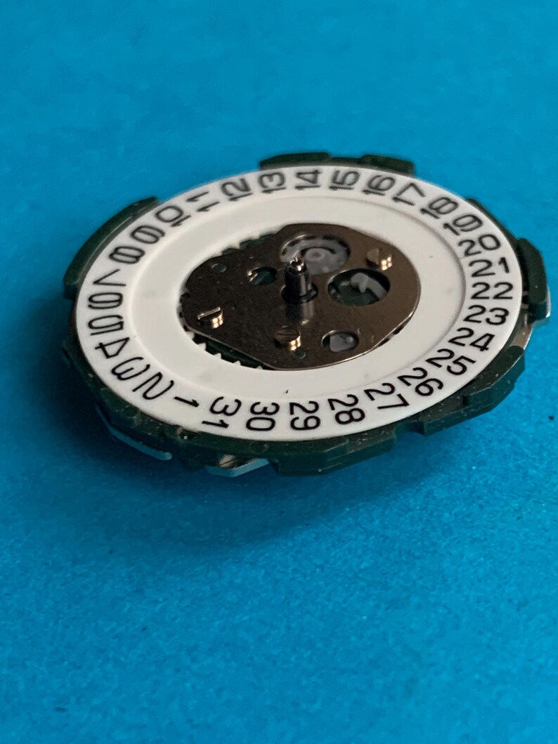 May include: A close-up of a watch movement, showing a white dial with black numbers for the days of the month. It has a green outer ring and a central mechanism with metallic parts. The background is a solid blue.