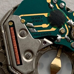 Puede incluir: Primer plano de un mecanismo de reloj con una placa de circuito verde y componentes met&aacute;licos. Los textos "SEVEN 7 JEWELS" y "UNADJUSTED" son visibles. Tambi&eacute;n hay una bobina de cobre y un volante dorado.
