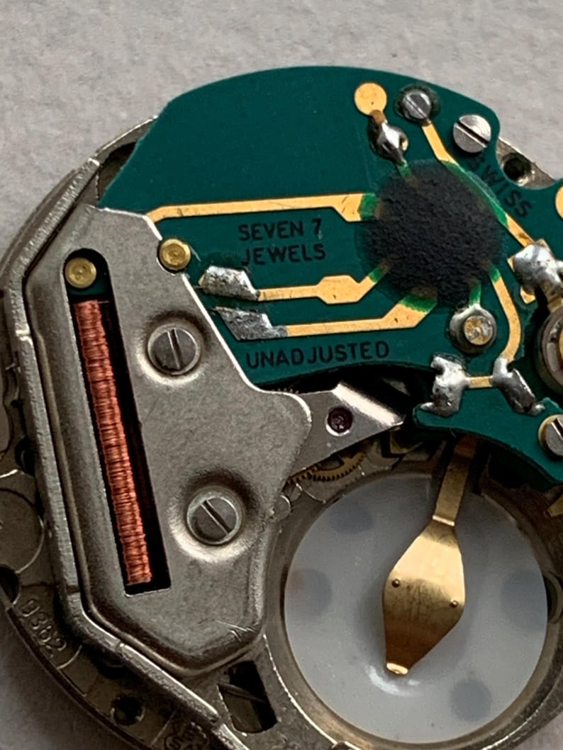 Puede incluir: Primer plano de un mecanismo de reloj con una placa de circuito verde y componentes met&aacute;licos. Los textos "SEVEN 7 JEWELS" y "UNADJUSTED" son visibles. Tambi&eacute;n hay una bobina de cobre y un volante dorado.