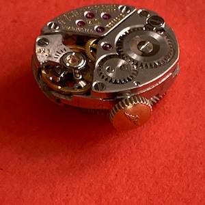 May include: Close-up of a vintage watch movement with intricate gears, jewels, and a gold-toned crown. The mechanical components are set against a red background, showcasing the detailed craftsmanship of the watch.