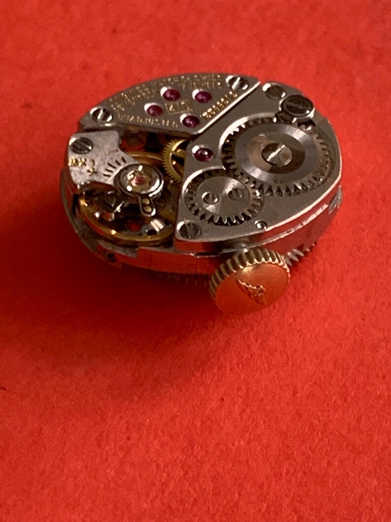 May include: Close-up of a vintage watch movement with intricate gears, jewels, and a gold-toned crown. The mechanical components are set against a red background, showcasing the detailed craftsmanship of the watch.