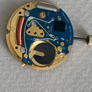 May include: Close-up of a watch movement with a blue circuit board and gold-colored metal components. The watch mechanism includes a battery, a copper coil, and the text "UNADJUSTED SEVEN 7 JEWELS".