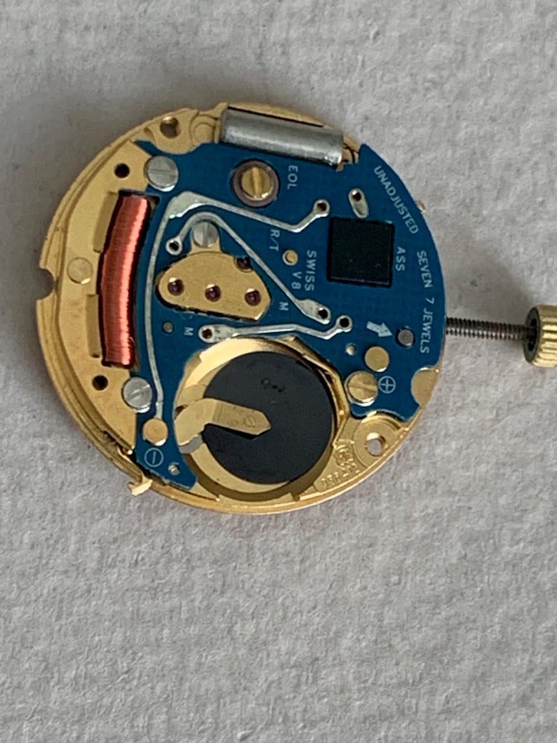 May include: Close-up of a watch movement with a blue circuit board and gold-colored metal components. The watch mechanism includes a battery, a copper coil, and the text "UNADJUSTED SEVEN 7 JEWELS".