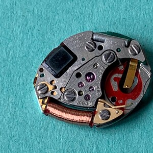 May include: Close-up of a watch movement with intricate gears, screws, and a red rotor. The watch mechanism is silver with gold accents and a small black square component. The text "SEIKO TIME CORP JAPAN" is visible.