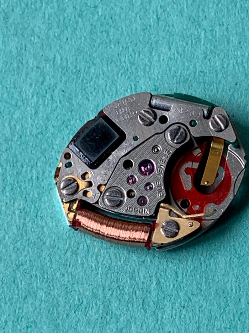 May include: Close-up of a watch movement with intricate gears, screws, and a red rotor. The watch mechanism is silver with gold accents and a small black square component. The text "SEIKO TIME CORP JAPAN" is visible.