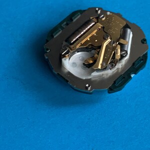 May include: A detailed view of a watch movement, featuring gold-coloured gears and various components. The mechanism is mounted on a silver base with a green surround. The background is a solid blue.