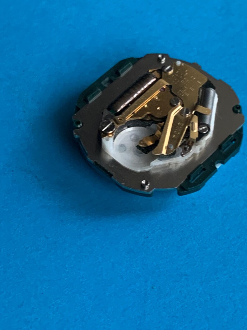 May include: A detailed view of a watch movement, featuring gold-coloured gears and various components. The mechanism is mounted on a silver base with a green surround. The background is a solid blue.