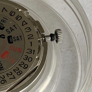 May include: Close-up of a watch movement with a silver-colored dial and a day-date display. The dial shows the days of the week and the date, with the date reading "6". A silver crown is visible on the side.