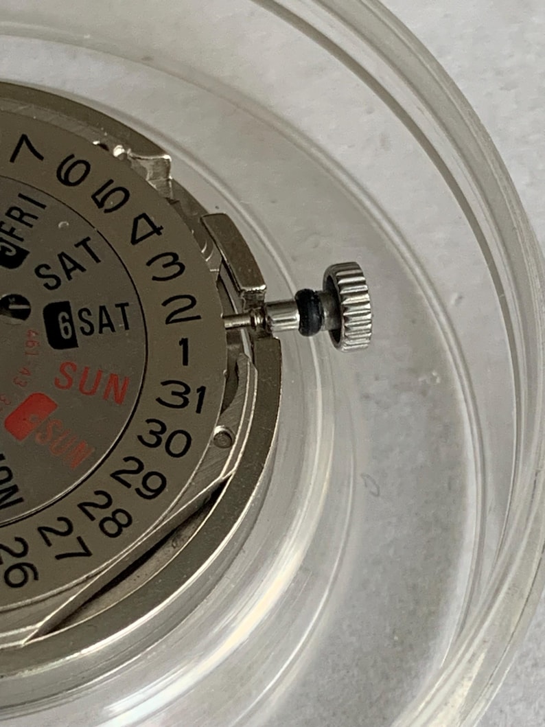 May include: Close-up of a watch movement with a silver-colored dial and a day-date display. The dial shows the days of the week and the date, with the date reading "6". A silver crown is visible on the side.