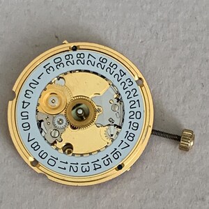 May include: A gold-toned watch movement with a light blue date dial and a screw-in crown. The dial displays the days of the month from 1 to 31. The watch mechanism is intricate and detailed.