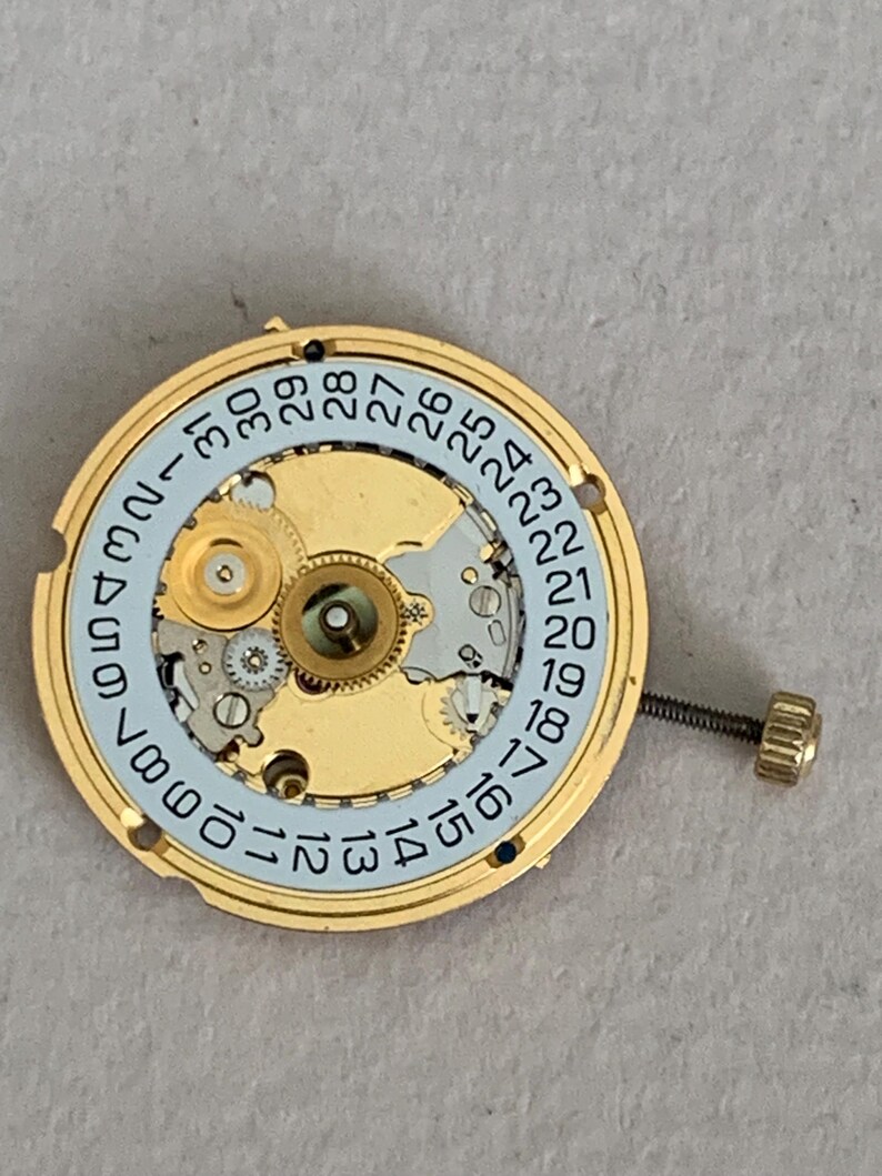 May include: A gold-toned watch movement with a light blue date dial and a screw-in crown. The dial displays the days of the month from 1 to 31. The watch mechanism is intricate and detailed.