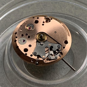 May include: Close-up of a watch movement with a copper-colored base and intricate gears. The mechanism includes silver-colored components and a small, protruding stem. The watch movement is set inside a clear, circular container.