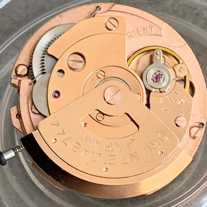 Puede incluir: Primer plano de un movimiento de reloj con una base de color cobre. El mecanismo presenta engranajes, tornillos y un volante con una joya rosa. El texto "ORIENT JAPAN 21J 4874" es visible.