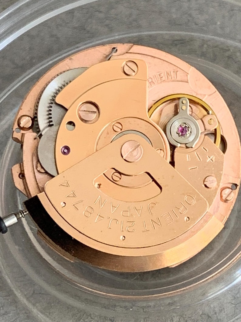Puede incluir: Primer plano de un movimiento de reloj con una base de color cobre. El mecanismo presenta engranajes, tornillos y un volante con una joya rosa. El texto "ORIENT JAPAN 21J 4874" es visible.