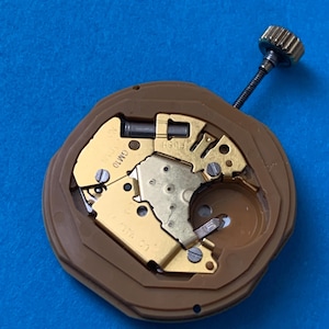 May include: A close-up of a watch movement with gold-colored gears and a brown plastic casing. The watch mechanism has a small crown and a stem. The background is a solid blue color.