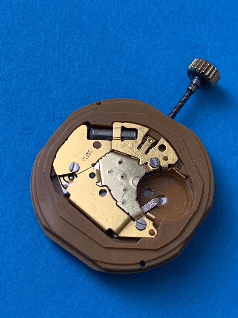 May include: A close-up of a watch movement with gold-colored gears and a brown plastic casing. The watch mechanism has a small crown and a stem. The background is a solid blue color.