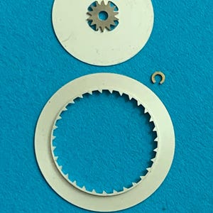 May include: A collection of watch repair parts, including a small metal gear, two white circular discs, and a tiny gold-colored C-clip. The discs have a toothed edge and a hole for the gear. The parts are arranged on a blue surface.