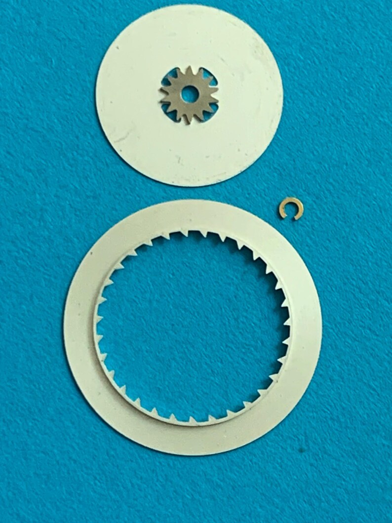 May include: A collection of watch repair parts, including a small metal gear, two white circular discs, and a tiny gold-colored C-clip. The discs have a toothed edge and a hole for the gear. The parts are arranged on a blue surface.