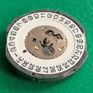 May include: A watch movement component with a white dial featuring black numerals from 1 to 31, indicating the days of the month. The component has a silver-colored outer ring and a central mechanism with small gears and screws.