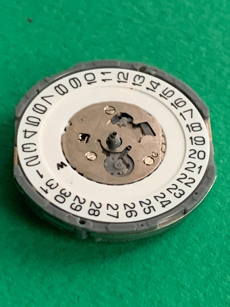 May include: A watch movement component with a white dial featuring black numerals from 1 to 31, indicating the days of the month. The component has a silver-colored outer ring and a central mechanism with small gears and screws.