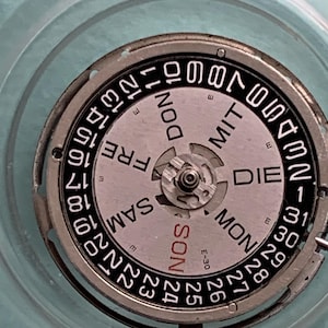 May include: Close-up of a watch movement with a silver and black dial. The dial displays the days of the week in German and numbers for the date. The watch movement is set against a light blue background.