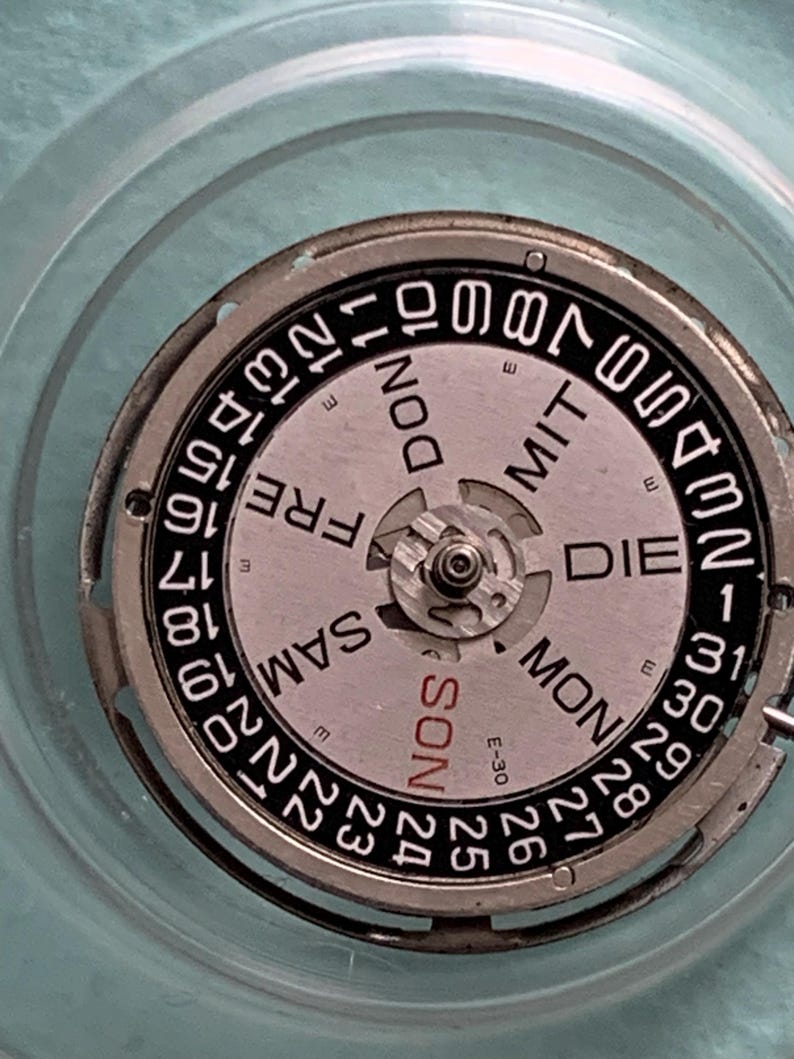 May include: Close-up of a watch movement with a silver and black dial. The dial displays the days of the week in German and numbers for the date. The watch movement is set against a light blue background.