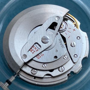 May include: Close-up of a watch movement with silver-coloured metal components. The mechanism includes gears, screws, and a small crown. The text "JAPAN" is visible on one of the parts. The overall design is intricate and detailed.