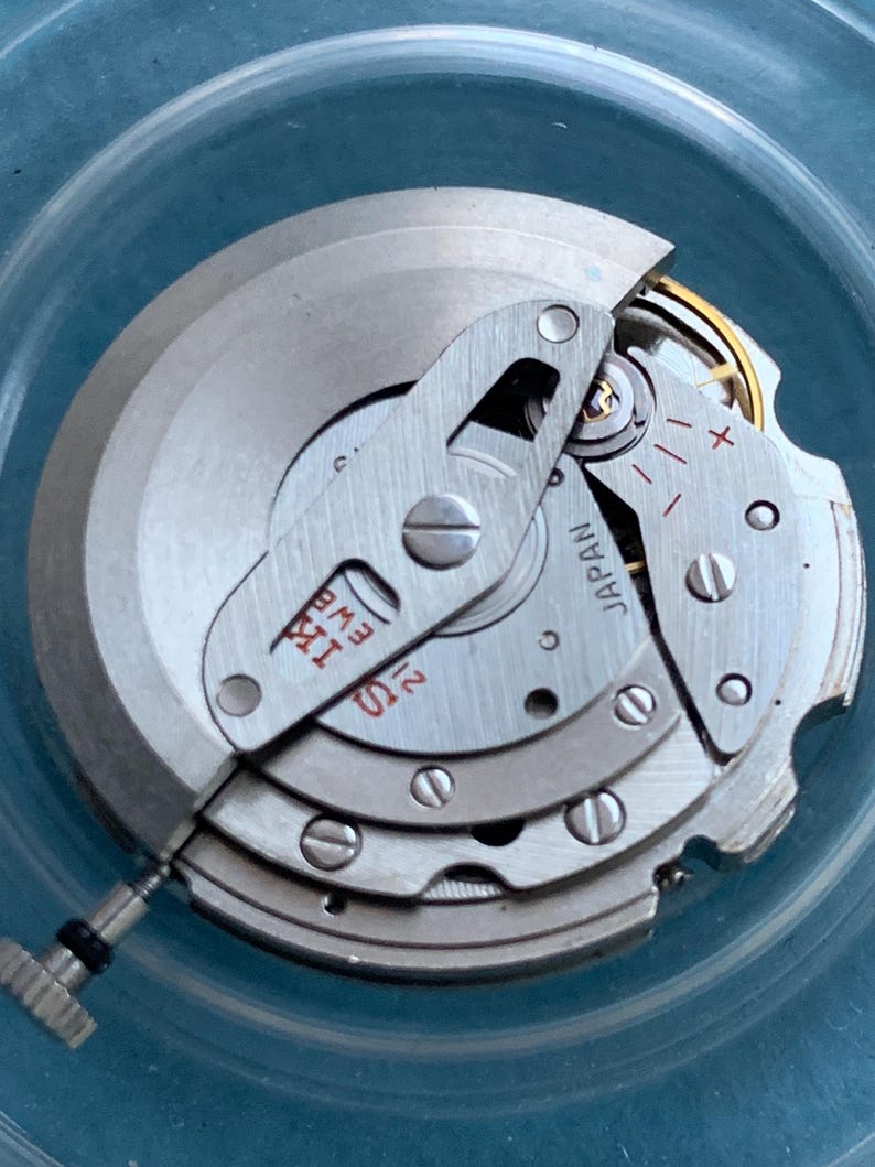 May include: Close-up of a watch movement with silver-coloured metal components. The mechanism includes gears, screws, and a small crown. The text "JAPAN" is visible on one of the parts. The overall design is intricate and detailed.