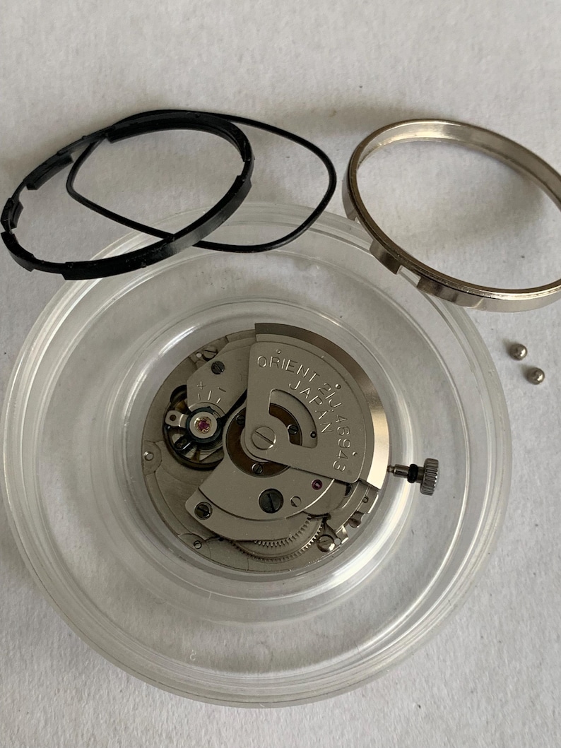 May include: A disassembled watch movement with various components. The main mechanism is silver-toned, marked "ORIENT JAPAN" and "46943".  Black rubber gaskets and a silver bezel ring are also present, along with small metal pins, all inside a clear plastic container.