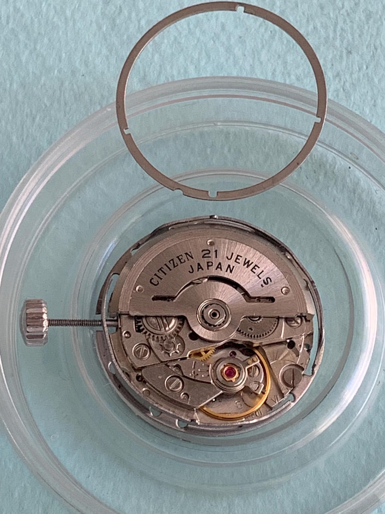 May include: A disassembled Citizen watch movement with the text "CITIZEN JAPAN 21 JEWELS" visible. The watch mechanism is silver-colored with intricate gears and a small red jewel. A silver-colored ring and a clear plastic holder surround the movement.