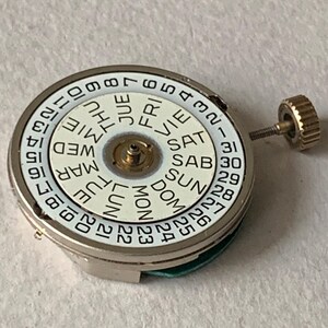 Puede incluir: Un movimiento de reloj con una esfera blanca que muestra los d&iacute;as de la semana y las fechas numeradas. La esfera est&aacute; rodeada por una caja de metal plateado. Una peque&ntilde;a corona dorada est&aacute; unida al lateral.