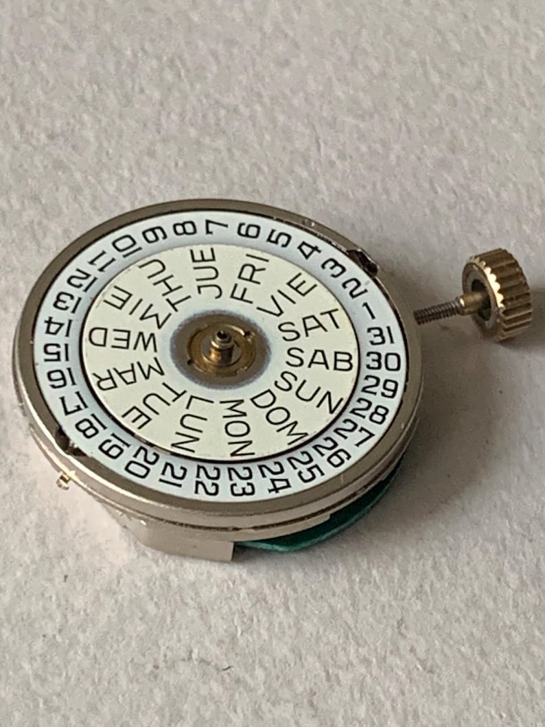 Puede incluir: Un movimiento de reloj con una esfera blanca que muestra los d&iacute;as de la semana y las fechas numeradas. La esfera est&aacute; rodeada por una caja de metal plateado. Una peque&ntilde;a corona dorada est&aacute; unida al lateral.