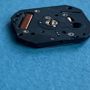 May include: Close-up of a black watch movement component with a copper coil and small gears. The component has several small holes and silver-colored screws. The background is a light blue surface.