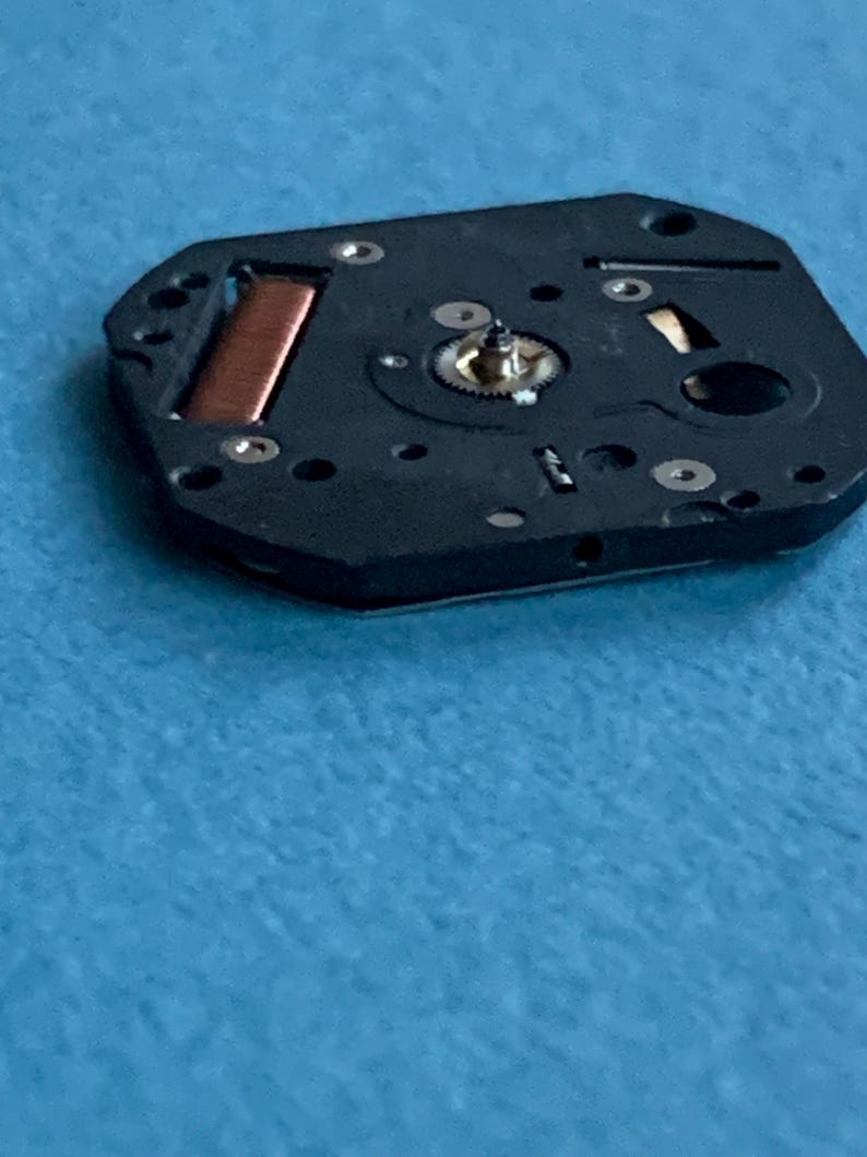 May include: Close-up of a black watch movement component with a copper coil and small gears. The component has several small holes and silver-colored screws. The background is a light blue surface.