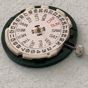 May include: A watch movement with a white dial displaying the days of the week in English and numbers 1-31. The dial is surrounded by a green plastic ring and has a silver winding stem.