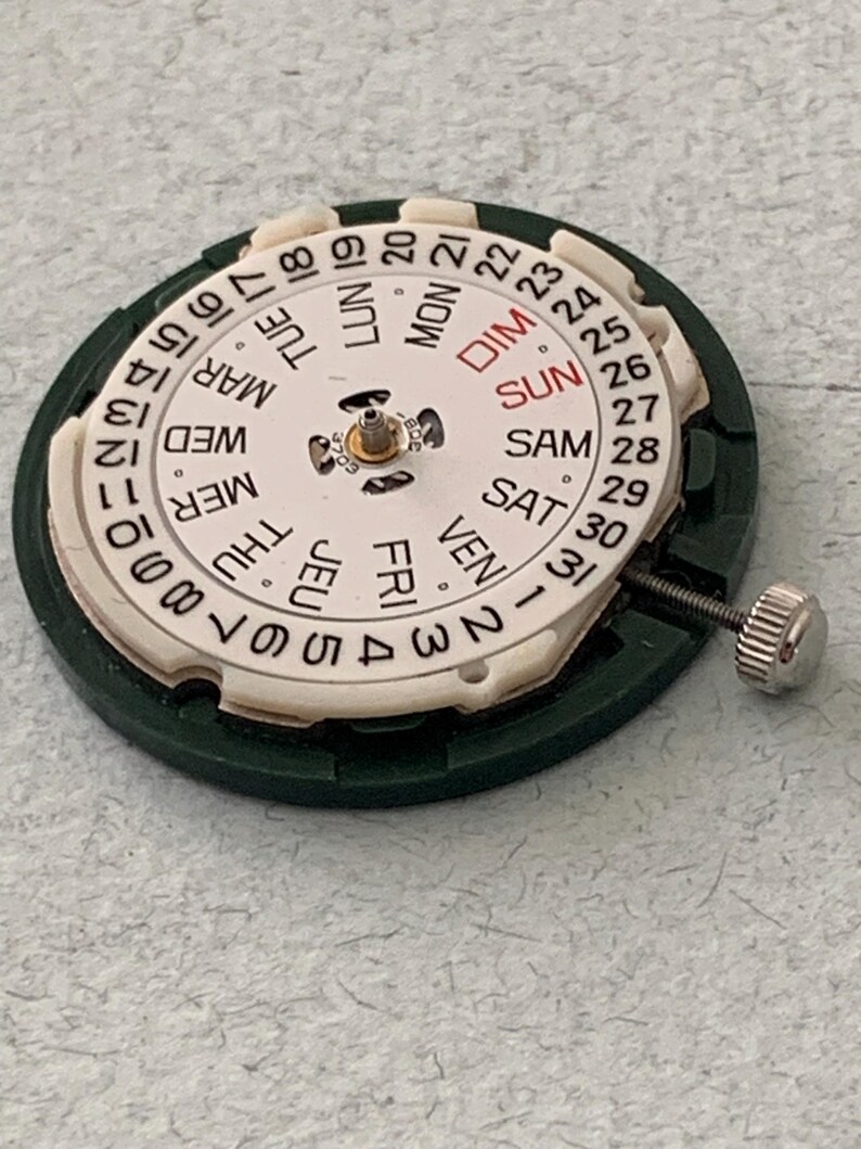 May include: A watch movement with a white dial displaying the days of the week in English and numbers 1-31. The dial is surrounded by a green plastic ring and has a silver winding stem.