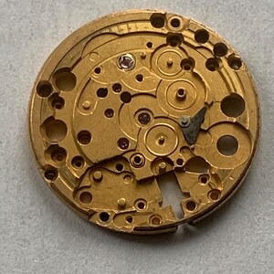 May include: Close-up of a gold-colored watch movement with intricate gears, holes, and small red jewels. The circular mechanism shows detailed craftsmanship, with various components and a complex design.