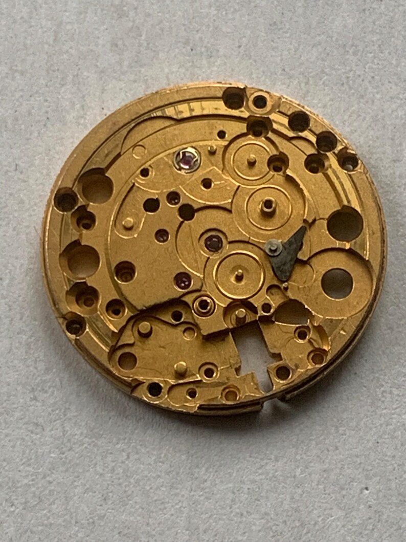 May include: Close-up of a gold-colored watch movement with intricate gears, holes, and small red jewels. The circular mechanism shows detailed craftsmanship, with various components and a complex design.