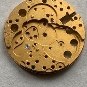 May include: A close-up of a gold-colored watch movement with intricate gears, holes, and small red jewels. The number "2571" is stamped on the edge. This mechanical component is likely for watch repair or restoration.