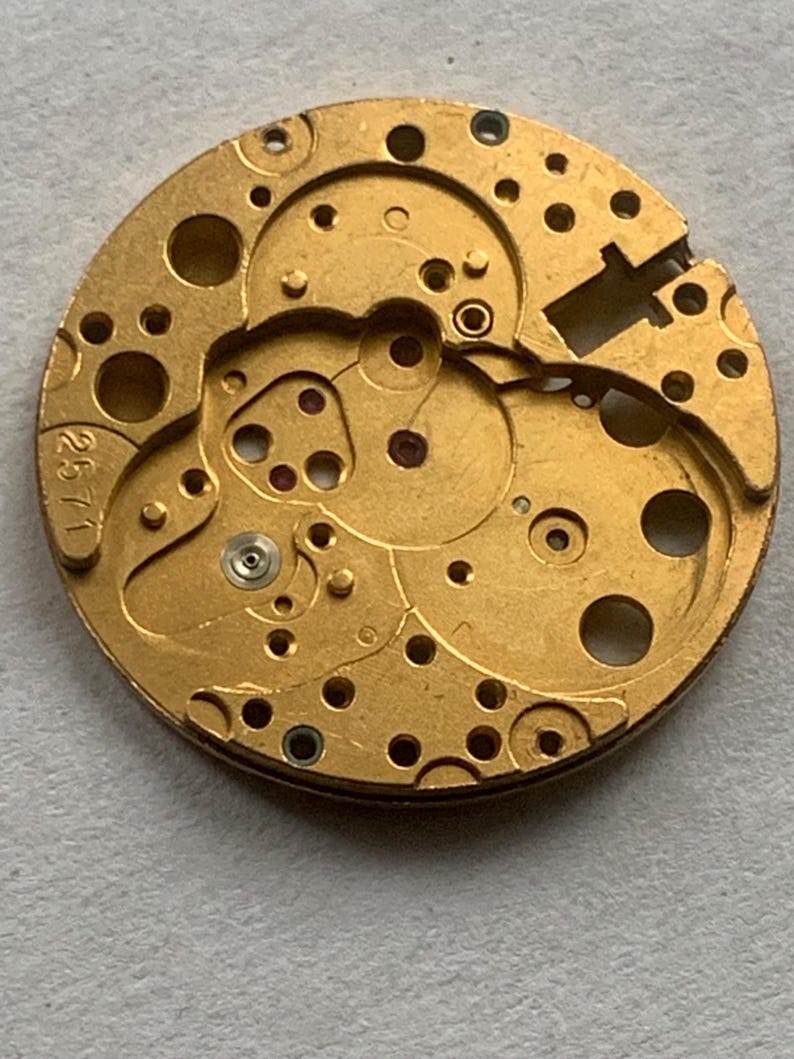 May include: A close-up of a gold-colored watch movement with intricate gears, holes, and small red jewels. The number "2571" is stamped on the edge. This mechanical component is likely for watch repair or restoration.