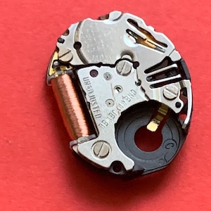 May include: Close-up of a watch movement with silver and gold components. The watch mechanism has a copper coil and the text "UNADJUSTED" and "ONE (1) JEWEL". The background is a solid red.