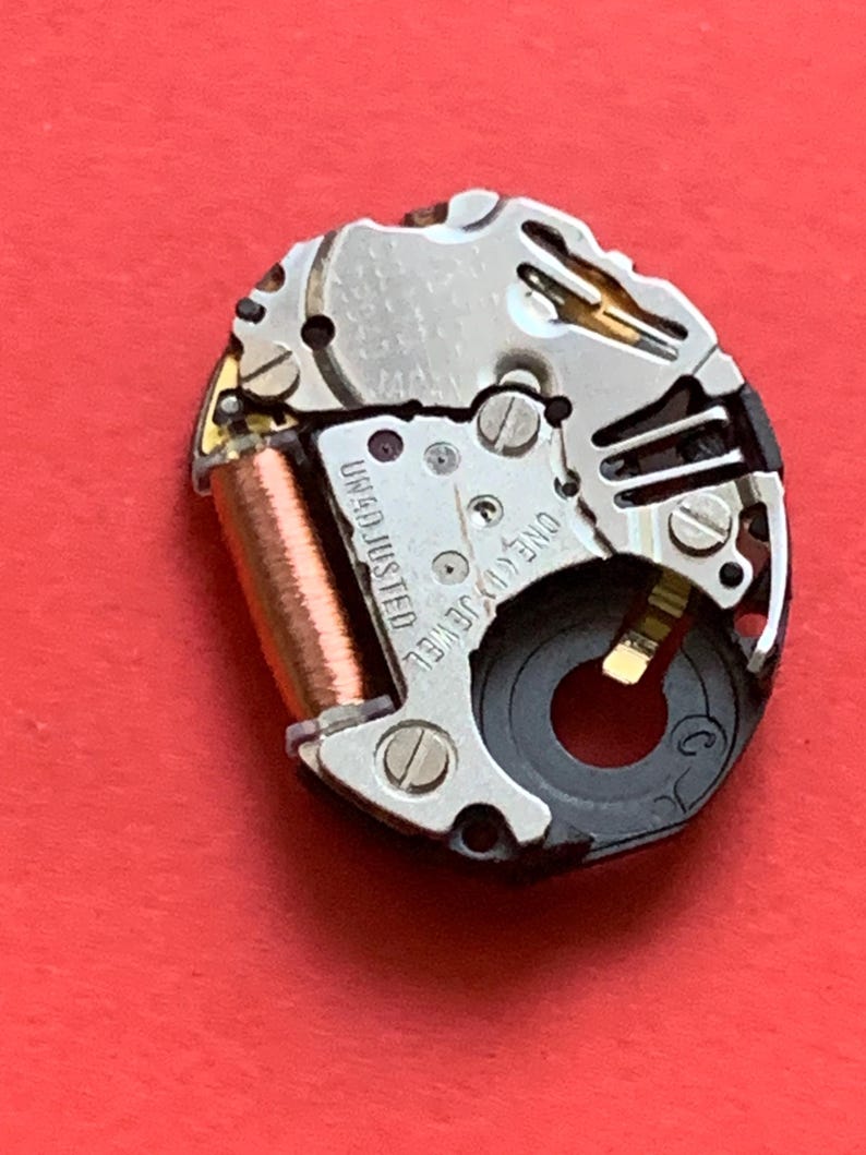 May include: Close-up of a watch movement with silver and gold components. The watch mechanism has a copper coil and the text "UNADJUSTED" and "ONE (1) JEWEL". The background is a solid red.