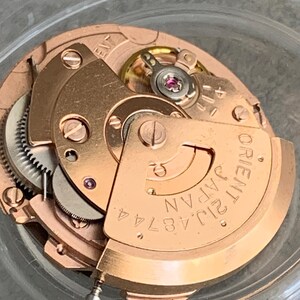 Puede incluir: Primer plano de un movimiento de reloj, mostrando engranajes y componentes intrincados. El movimiento es de color cobre, con la inscripci&oacute;n "ORIENT JAPAN 21J 48744 A" en una placa curva. La imagen resalta la precisi&oacute;n del mecanismo del reloj.