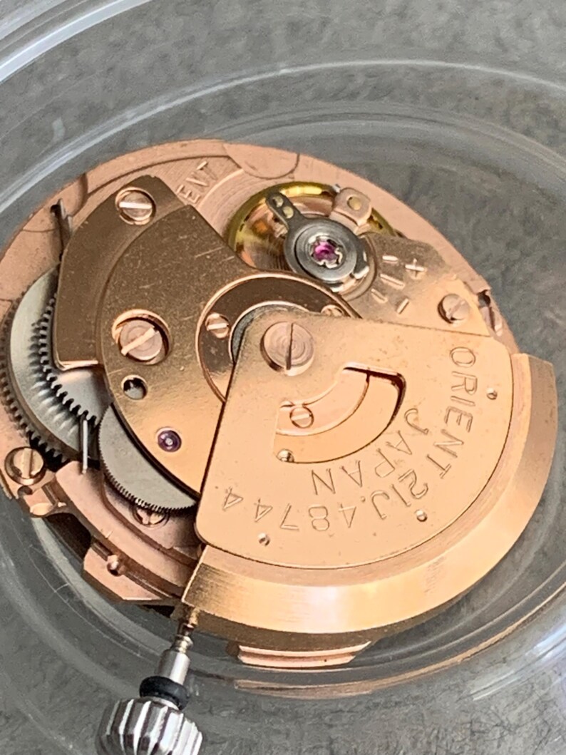 Puede incluir: Primer plano de un movimiento de reloj, mostrando engranajes y componentes intrincados. El movimiento es de color cobre, con la inscripci&oacute;n "ORIENT JAPAN 21J 48744 A" en una placa curva. La imagen resalta la precisi&oacute;n del mecanismo del reloj.