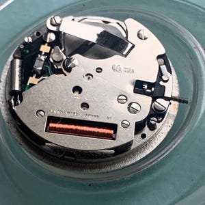 Op de afbeelding: Close-up van een uurwerk met zilveren en goudkleurige componenten. Het mechanisme heeft kleine schroeven, tandwielen en een rode rechthoekige spoel. De tekst "FIVE JEWELS" en "UNADJUSTED SWISS" is zichtbaar.