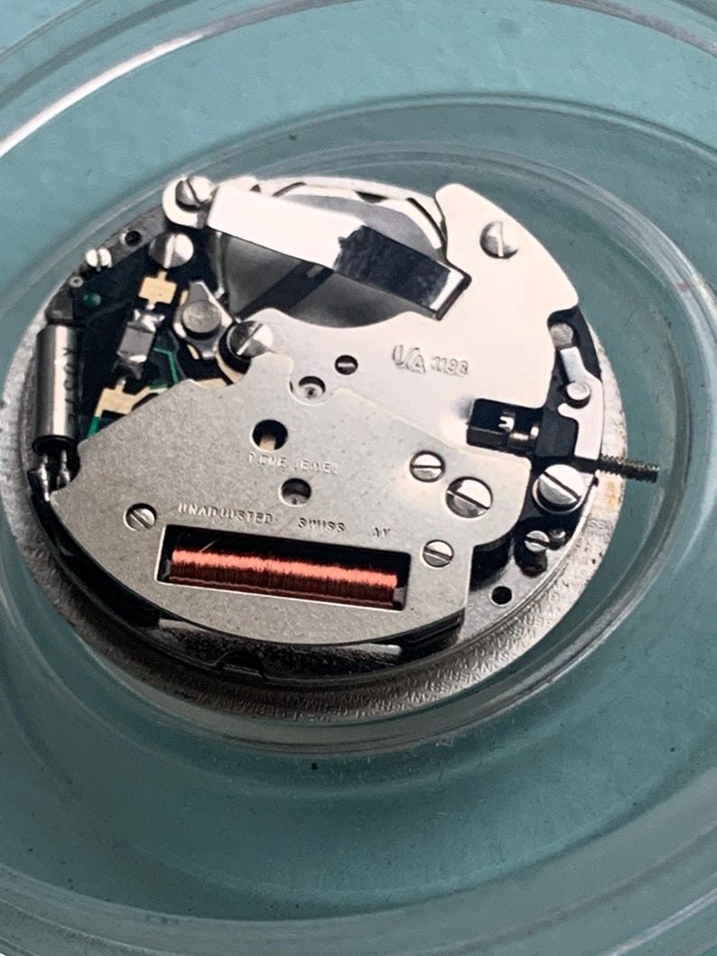 Op de afbeelding: Close-up van een uurwerk met zilveren en goudkleurige componenten. Het mechanisme heeft kleine schroeven, tandwielen en een rode rechthoekige spoel. De tekst "FIVE JEWELS" en "UNADJUSTED SWISS" is zichtbaar.